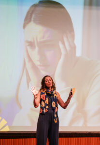 Stephanie Krauss on stage infront of a screen with a student holding her head
